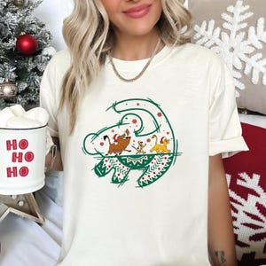 May include: Cream-colored t-shirt featuring a green and red Christmas-themed graphic of characters from The Lion King. The graphic includes Simba, Timon, and Pumbaa wearing Santa hats. The shirt is a relaxed fit.
