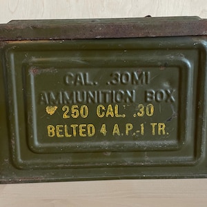 May include: An olive green ammunition box with a hinged lid and a metal handle. The box has yellow lettering that reads "CAL .30MI AMMUNITION BOX 250 CAL .30 BELTED 4 A.P.-1 TR."