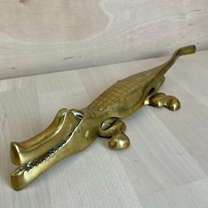 May include: A brass nutcracker shaped like an alligator with a textured back, a hinged jaw mechanism, and four short legs. The metal has a polished gold-toned finish with visible detailing on the snout and body, designed to open when the tail is lifted.