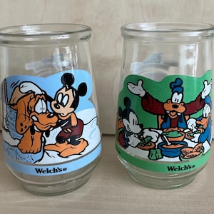 May include: Two clear glass Welch's jars with cartoon character designs. One jar features Mickey Mouse and Pluto on a blue background, the other features Mickey, Goofy, and Donald Duck on a green background. The text "Welch's" is printed on the bottom.