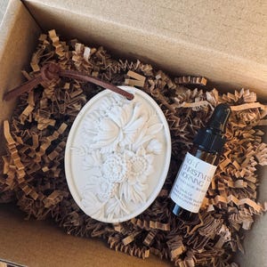 May include: A white, oval floral ornament with a brown leather cord, presented in a cardboard box with brown paper. A small bottle with a black dropper and a label reading "No. 1 Christmas Morning" is also in the box.