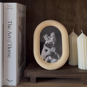 Peut inclure: Un cadre photo ovale de couleur neutre avec une photo en noir et blanc d'une mère et d'un enfant. Deux bougies blanches et deux livres sont sur une étagère en bois foncé. Les livres sont intitulés "design select/ion" et "The Art of Home."