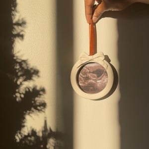 May include: A white, circular ornament with a bow detail at the top, displaying a sonogram image. It is suspended by a rust-coloured ribbon. The ornament is held by a person's hand against a cream-coloured wall.