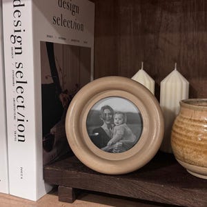 May include: A round, light-colored wooden picture frame with a black and white photo of a mother and child. The frame sits on a dark wooden shelf with books and candles. The text "THE NOVA COLLECTION" is visible.