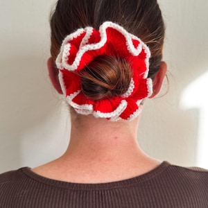 Peut inclure: Un chouchou rouge et blanc au crochet, en forme de fleur, est porté dans un chignon. Le chouchou a un bord à volants et est fait de fil rouge avec une bordure blanche. La personne porte un haut côtelé marron.