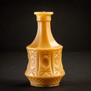 Puede incluir: Una vela decorativa de color ámbar con forma de jarrón. La vela presenta intrincados patrones de estrellas y flores tallados en su superficie. La vela tiene una mecha y se encuentra sobre un fondo oscuro.