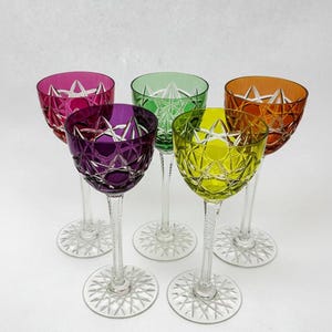 May include: Five colorful crystal wine glasses. The glasses have a starburst pattern on the base and a diamond pattern on the bowl. The colors are pink, purple, green, yellow, and orange. The stems are clear.