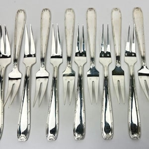 May include: A collection of ten silver-colored forks arranged on a light background. Each fork has a slender handle with decorative detailing and three tines. The forks are evenly spaced, showcasing their design and craftsmanship.