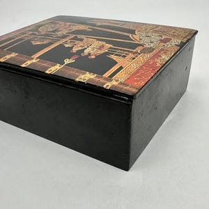 May include: A rectangular black box with a decorative lid. The lid features a detailed, colorful design with gold, brown, and red accents. The box appears to be made of wood and has a simple, elegant design.