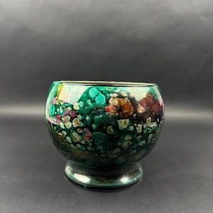 Peut inclure: Un bol décoratif en verre de forme arrondie, avec un petit socle. Le bol présente un motif tacheté vibrant avec des nuances de vert, de marron et de rouge. L'intérieur est visible, et l'effet global est artistique et coloré.