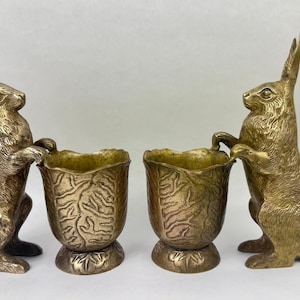 May include: A pair of brass rabbit figurines standing on their hind legs, each holding a textured, leaf-shaped vessel. The rabbits have detailed fur textures and pointed ears, while the attached containers feature a ribbed, organic surface.