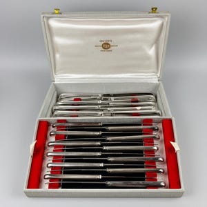 May include: A set of silver-colored knives in a gray box with a red velvet interior. The box is open, revealing the knives neatly arranged. The top of the box has a satin lining and gold-colored clasps. The text "COUVERTS STAR" is visible on the inside of the lid.