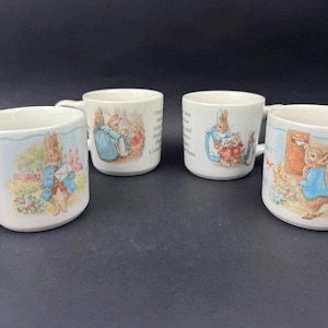 Może przedstawiać: Cztery białe ceramiczne kubki z ilustracjami królika Piotrusia w niebieskiej kurtce. Każdy kubek przedstawia inne sceny z opowieści, w tym królika Piotrusia z kopertą, w pobliżu ogrodu oraz z innymi królikami, wraz z nadrukowanym tekstem.