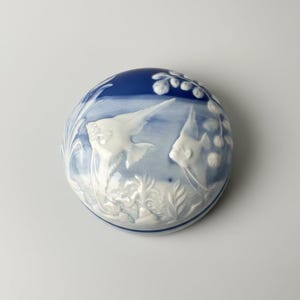 Peut inclure: Une boîte à bijoux ronde en porcelaine avec une palette de couleurs bleu profond et blanc. Le couvercle présente un motif en relief de deux poissons-anges nageant parmi des plantes aquatiques et des bulles, avec une finition blanche lisse.