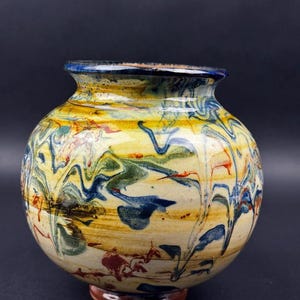 May include: A round ceramic vase with a wide body and a narrow neck. The vase features a colorful, swirled design with yellow, blue, green, and red hues. The rim is blue, and the base is a dark brown color.