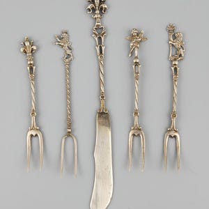 Op de afbeelding: Een set van vijf zilverkleurige metalen bestekstukken, bestaande uit vier vorken met twee tanden en één mes met een plat lemmet. Elk handvat is voorzien van een unieke decoratieve knop, waaronder een fleur-de-lis, een leeuw, een cherubijn en een leeuw met een kroon.