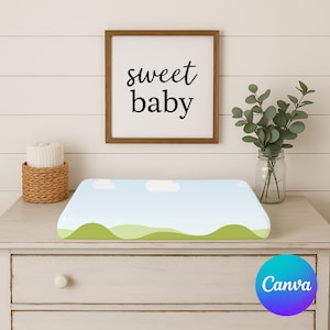 May include: A baby changing mat with a landscape design featuring a blue sky, white clouds, and green hills. A framed sign above reads "sweet baby". A small basket with a rolled-up towel and a glass jar with greenery are also visible.