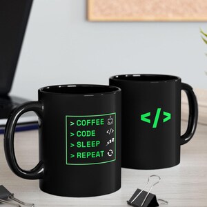 K&ouml;nnte beinhalten: Zwei schwarze Kaffeetassen mit wei&szlig;em Text und gr&uuml;nen Akzenten. Eine Tasse hat den Text "COFFEE > CODE > SLEEP > REPEAT" mit Symbolen. Die andere Tasse hat ein gr&uuml;nes Codesymbol. Die Tassen stehen auf einer hellen Oberfl&auml;che.