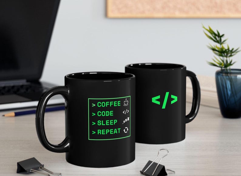K&ouml;nnte beinhalten: Zwei schwarze Kaffeetassen mit wei&szlig;em Text und gr&uuml;nen Akzenten. Eine Tasse hat den Text "COFFEE > CODE > SLEEP > REPEAT" mit Symbolen. Die andere Tasse hat ein gr&uuml;nes Codesymbol. Die Tassen stehen auf einer hellen Oberfl&auml;che.