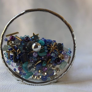 Pu&ograve; includere: Una sfera di vetro con bordo argentato piena di una variet&agrave; di perline colorate, tra cui blu, turchese, viola e oro. Le perline sono disposte in uno schema casuale, creando un design visivamente accattivante e intricato.