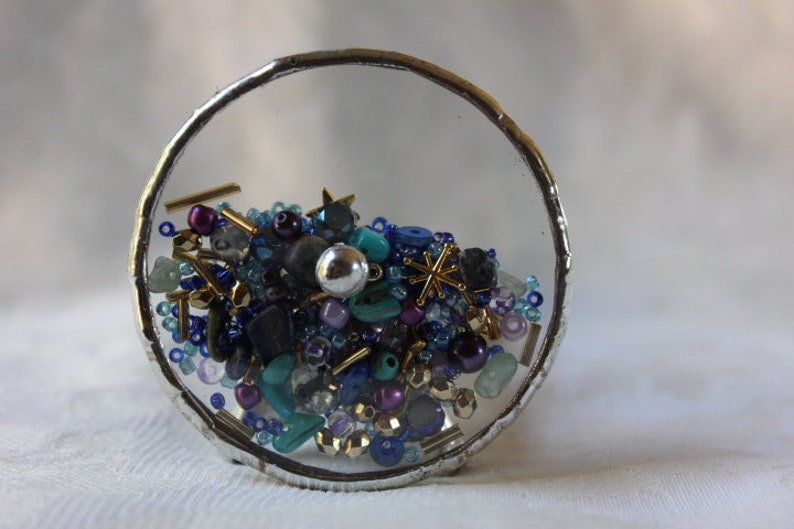 Pu&ograve; includere: Una sfera di vetro con bordo argentato piena di una variet&agrave; di perline colorate, tra cui blu, turchese, viola e oro. Le perline sono disposte in uno schema casuale, creando un design visivamente accattivante e intricato.