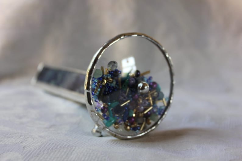 Pu&ograve; includere: Un caleidoscopio argentato con un cilindro di vetro trasparente pieno di perline colorate e piccoli pezzi di vetro. Le perline sono blu, viola, verdi e dorate.