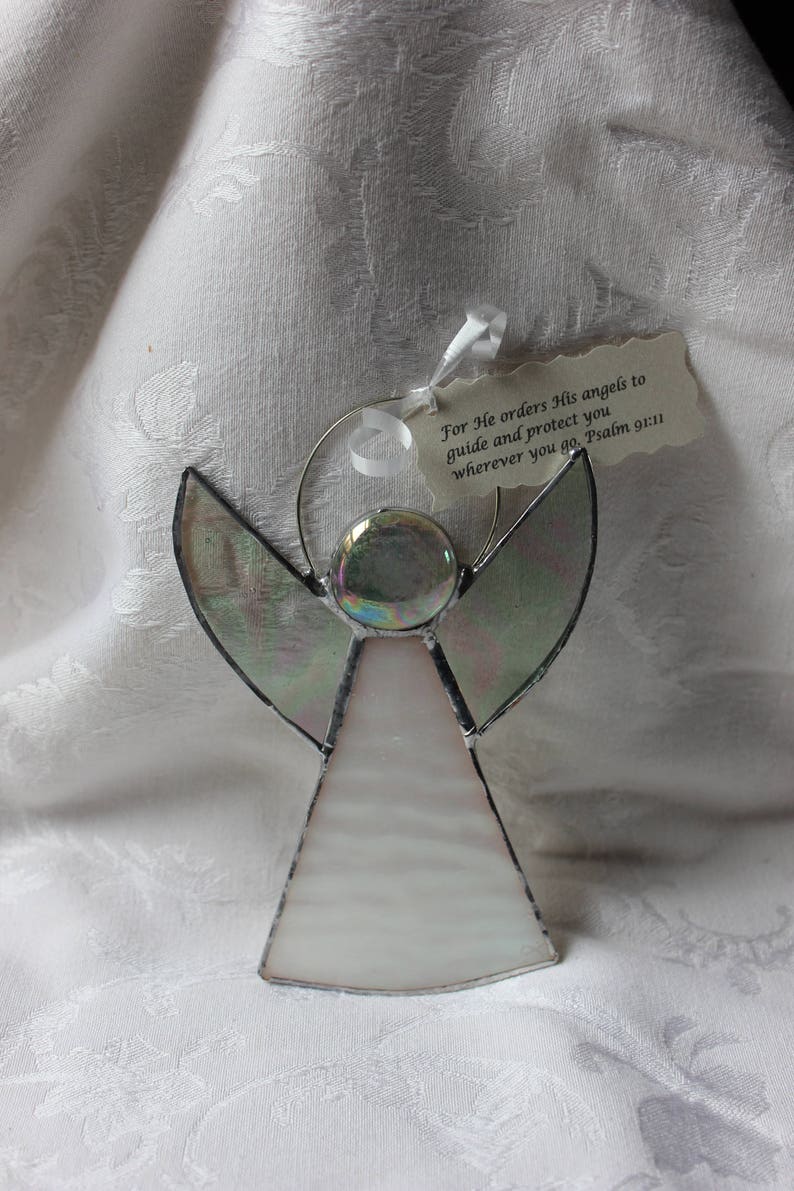 May include: A stained glass angel ornament with iridescent wings and a white body. The ornament has a silver wire loop for hanging and a tag with the text "For He orders His angels to guide and protect you wherever you go. Psalm 91:11".