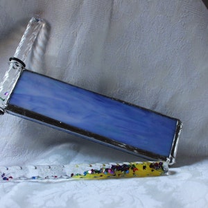 May include: A handmade kaleidoscope with a rectangular blue glass panel and clear, glitter-filled handles. One handle is pink, the other is yellow. The kaleidoscope is set against a white, textured background.