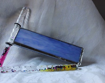May include: A handmade kaleidoscope with a rectangular blue glass panel and clear, glitter-filled handles. One handle is pink, the other is yellow. The kaleidoscope is set against a white, textured background.