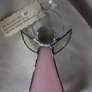 May include: A stained glass angel ornament with a pink body and clear wings. The angel is holding a clear glass circle in its hands. The ornament is hanging from a silver wire. The text on the tag reads "For He orders His angels guide and protect you. Wherever you go. Psalm 91:11"
