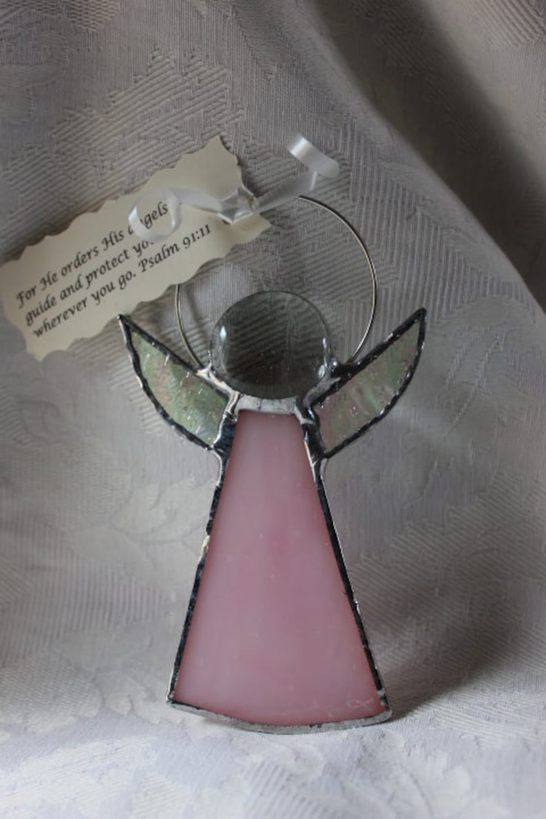May include: A stained glass angel ornament with a pink body and clear wings. The angel is holding a clear glass circle in its hands. The ornament is hanging from a silver wire. The text on the tag reads "For He orders His angels guide and protect you. Wherever you go. Psalm 91:11"