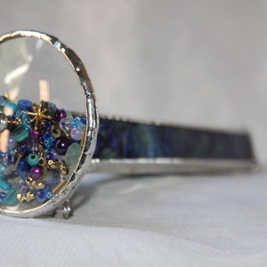 Pu&ograve; includere: Un caleidoscopio argentato con un cilindro di vetro trasparente pieno di perline colorate e una base di vetro blu e verde.