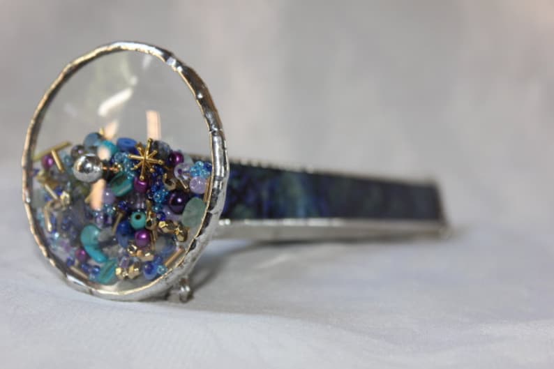 Pu&ograve; includere: Un caleidoscopio argentato con un cilindro di vetro trasparente pieno di perline colorate e una base di vetro blu e verde.