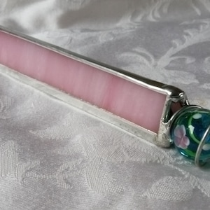 May include: A handmade kaleidoscope with a pink glass body and a silver-colored metal frame. The end features a decorative glass ball with a floral design in shades of blue, green, and pink. The kaleidoscope is designed for viewing patterns.