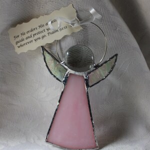 May include: Pink stained glass angel ornament with a clear glass center and silver wire halo. The ornament has a tag with the text "For He orders His angels guide and protect you wherever you go. Psalm 91:11".