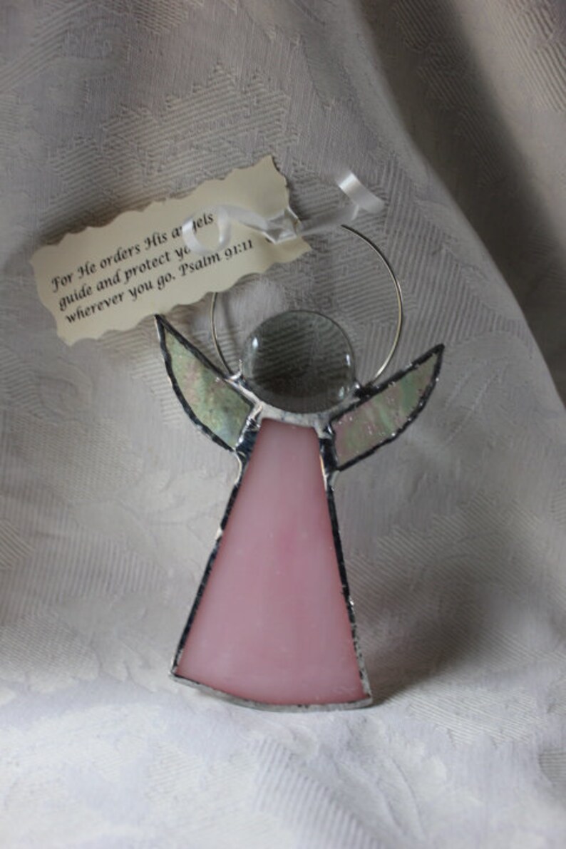 May include: Pink stained glass angel ornament with a clear glass center and silver wire halo. The ornament has a tag with the text "For He orders His angels guide and protect you wherever you go. Psalm 91:11".