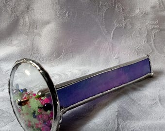 May include: A kaleidoscope with a clear glass end and a purple and pink stained glass body. The kaleidoscope is filled with colorful beads and sequins.