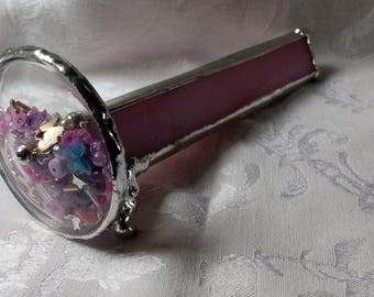 May include: A kaleidoscope constructed with pink glass panels and silver solder joints. The circular viewing end features a clear glass window containing a collection of loose, colorful beads, small butterfly shapes, and metallic accents.