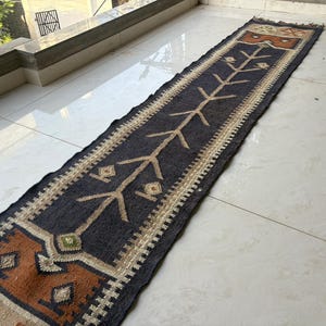 May include: A long, rectangular rug with a dark blue central field and geometric patterns. The rug features brown, beige, and green accents, with a repeating tree-like design in the centre. The edges are finished with a beige border.