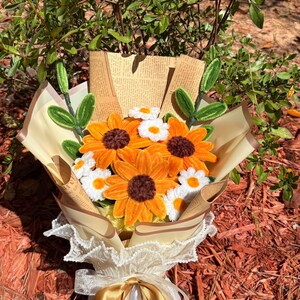 May include: A bouquet of handmade sunflowers and daisies wrapped in tan paper and lace. The sunflowers have orange petals and brown centers, while the daisies are white with yellow centers. The bouquet is tied with a gold ribbon.