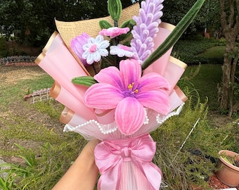 May include: A bouquet of handcrafted pipe cleaner flowers featuring a large pink lily, lavender-colored spikes, and small white daisies with green stems. The arrangement is wrapped in pink and tan paper with a large pink ribbon tied at the base.