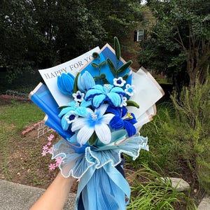 May include: A bouquet of handmade blue and white crochet flowers, including lilies and tulips, wrapped in blue paper and tulle. The bouquet has a ribbon and the text "HAPPY FOR YOU".