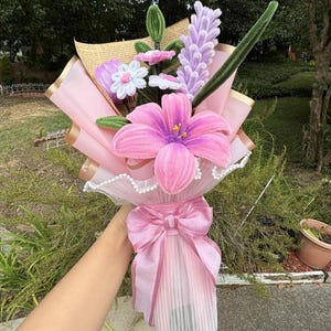 May include: A handmade bouquet of fabric flowers in shades of pink, purple, and white. The bouquet includes a large pink lily, smaller flowers, and a lavender-colored stem. The bouquet is wrapped in pink paper and tied with a pink bow.