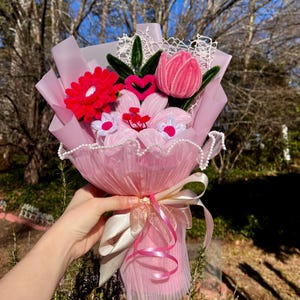 May include: A handmade bouquet of crocheted flowers in shades of pink and red, wrapped in pink paper and tied with a cream ribbon. The bouquet includes a red daisy, a pink tulip, and other floral elements. The background shows a blurred outdoor scene.
