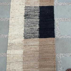 May include: A rectangular woven rug with a geometric pattern. The rug features alternating blocks of beige, tan, black, and white. The rug is made of a natural fiber material, and the texture is visible. The rug is placed on a gray stone surface.
