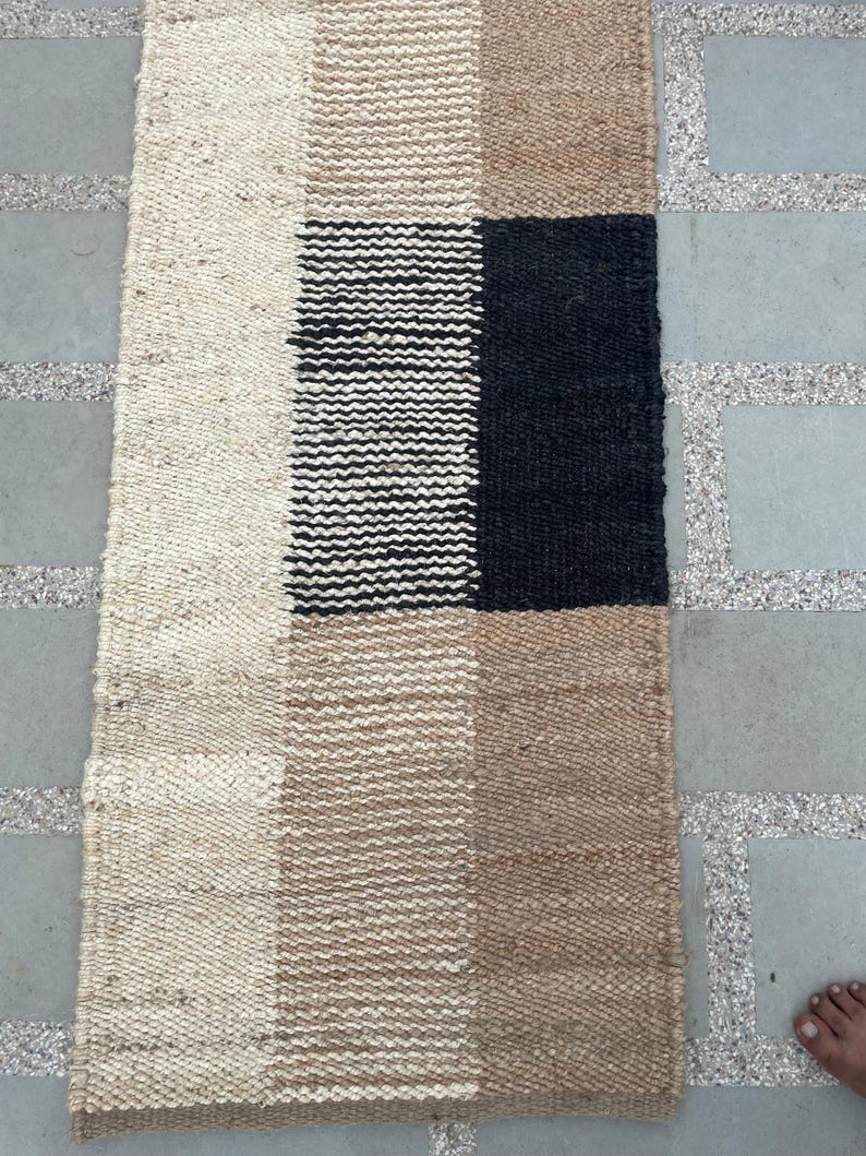 May include: A rectangular woven rug with a geometric pattern. The rug features alternating blocks of beige, tan, black, and white. The rug is made of a natural fiber material, and the texture is visible. The rug is placed on a gray stone surface.