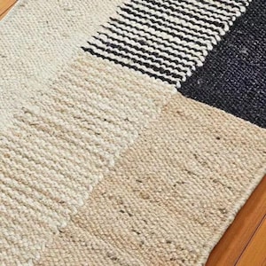 May include: A rectangular rug with a geometric design. The rug features sections of cream, tan, black and navy blue. The rug is made of a woven material with a textured surface.
