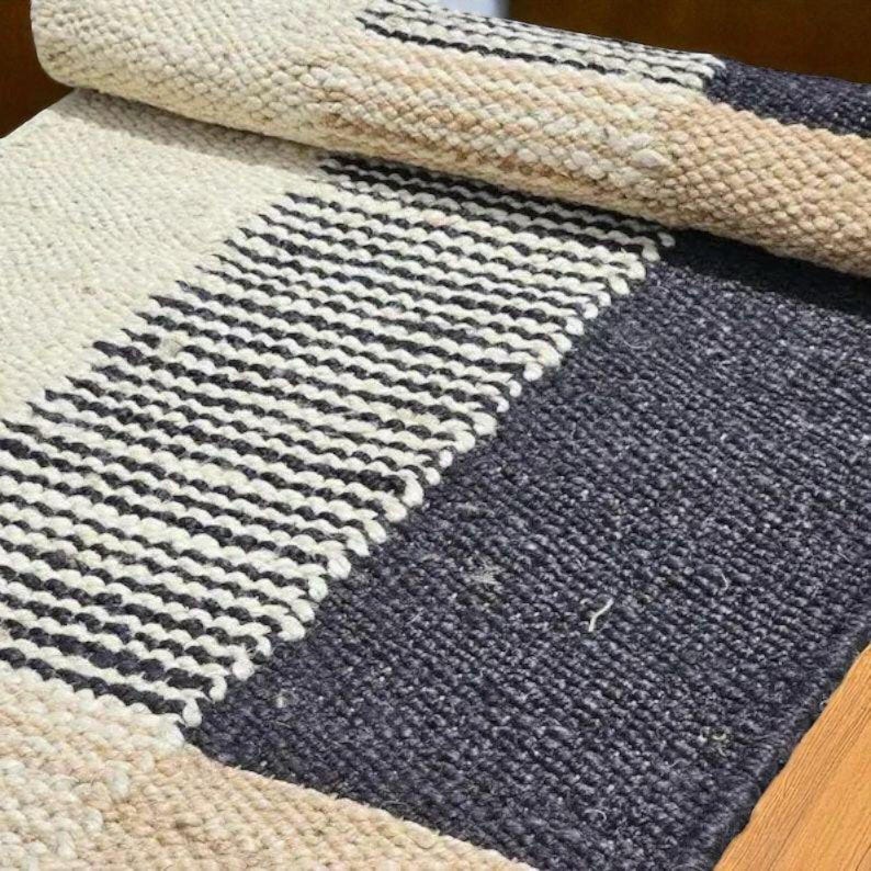 May include: A rectangular woven rug with a striped pattern. The rug features alternating bands of off-white, beige, black and navy blue. The rug is partially rolled up, revealing the underside and the texture of the weave.