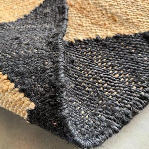 May include: Close-up of a woven rug with a geometric pattern. The rug features a combination of black and tan sections, creating a textured surface. The edges of the rug are visible, showcasing the woven construction.