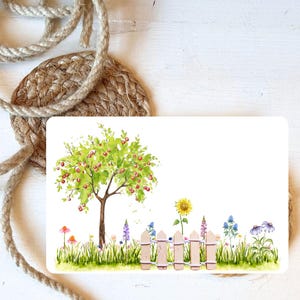 Könnte beinhalten: Eine rechteckige Karte mit einer Aquarellillustration eines Baumes mit roten Früchten, einem Holzzaun und verschiedenen Blumen. Die Karte liegt auf einem gewebten Untersetzer und einem Seil.